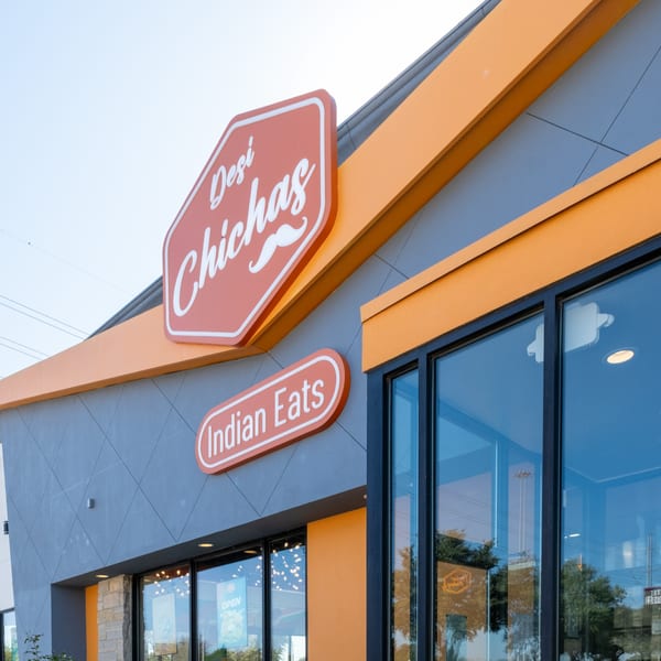 Exterior of Desi Chichas Indian Eats restaurant with orange and gray facade and large windows.