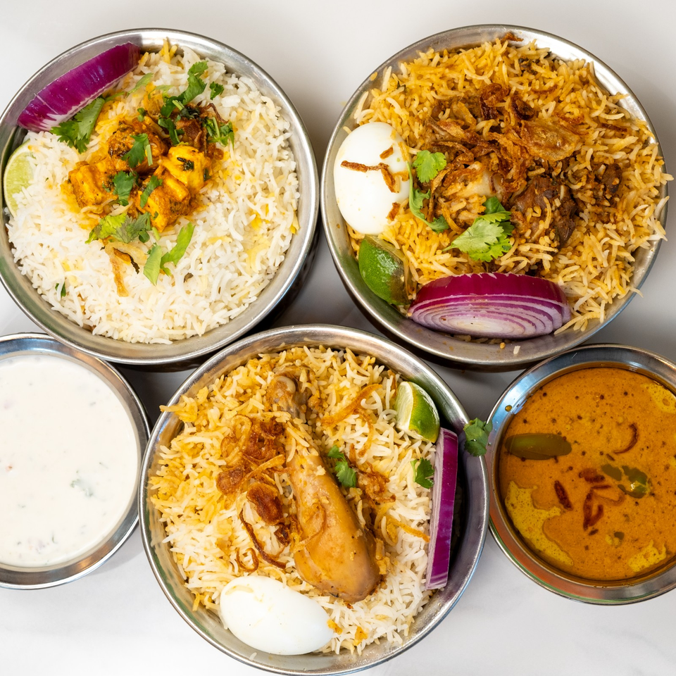 Overhead spread of Indian biryani bowls with chicken, eggs, cilantro, onions, lime, raita, and curry.