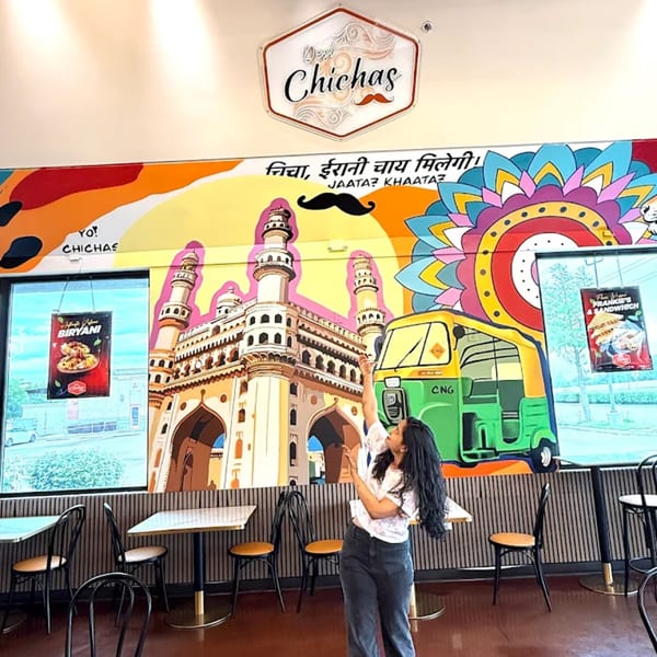 A woman poses in front of a colorful mural depicting Indian landmarks and an auto-rickshaw at Desi Chichas.