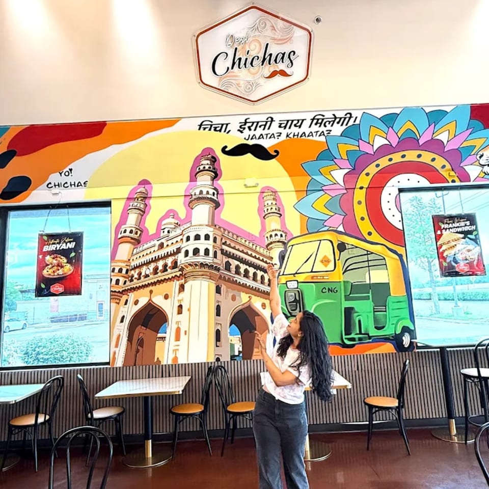 A woman poses in front of a colorful mural depicting Indian landmarks and an auto-rickshaw at Desi Chichas.