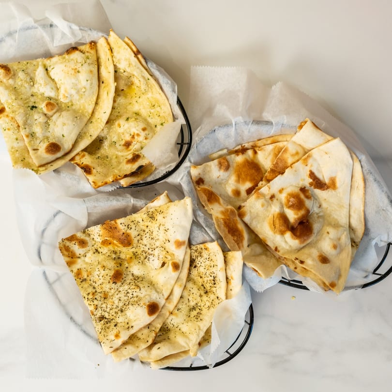 Three baskets of Indian naan bread, some topped with herbs, on a white marble surface.