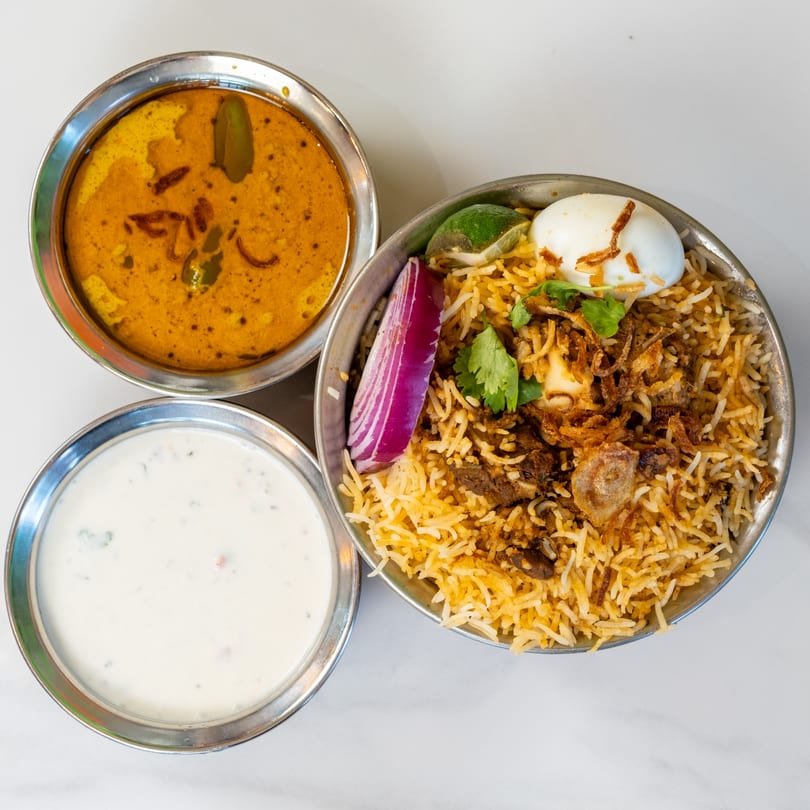 Indian biryani with egg, red onion, and lime, served with curry and raita in steel bowls.