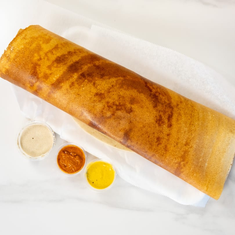 A long, golden-brown dosa from Desi Chichas, served with three small cups of dipping sauces.