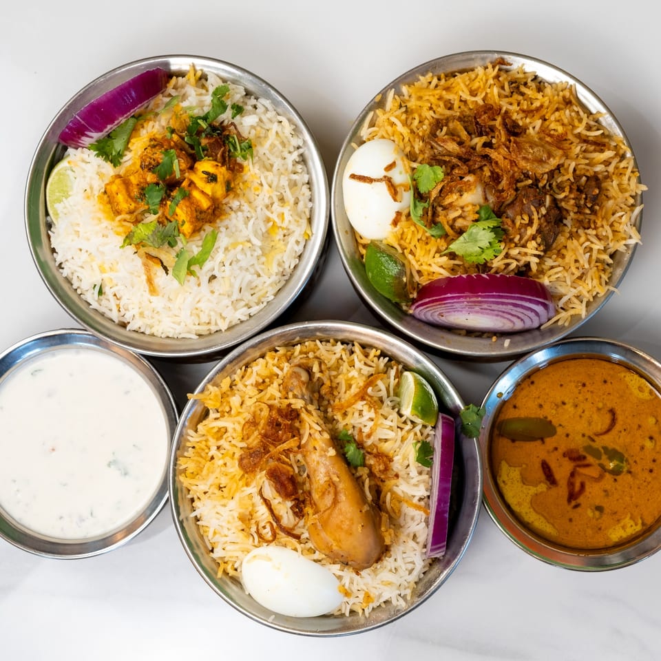 Four bowls of Indian biryani, two with chicken, one with paneer, and one with mutton, plus a side of raita and curry.