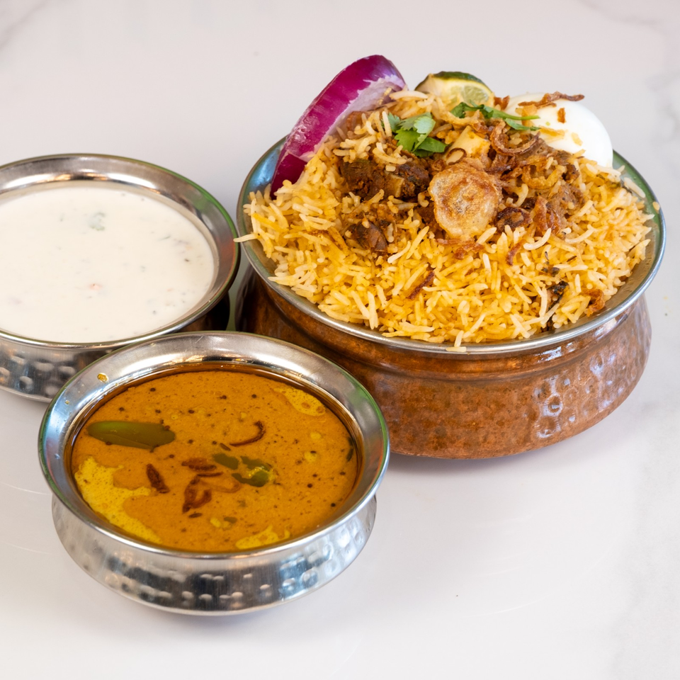 Indian biryani with fried onions, cilantro, and egg served with creamy raita and spiced curry in metal bowls.